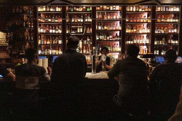 A row of customers sit at a bar with a wall of bottles behind it. A bartender pours a beer from a tap.